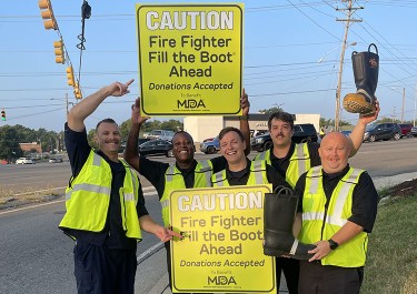 firefighters stand on a sidewalk holding signs promoting Fill the Boot