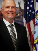 Man with a goatee in a suit standing next to flags.